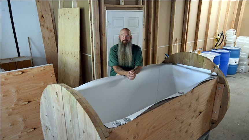 Man (Seth Viddal) leans on his wooden body composting 'vessel' in a warehouse. He has a long beard.
