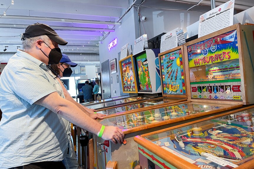 Гости посещают Silverball Retro Arcade, где мужчина играет в автомат для игры в пинбол в маске.