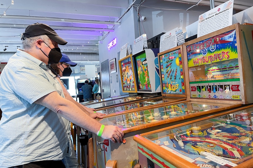 Guests visit Silverball Retro Arcade,  man plays pinball machine wearing face mask