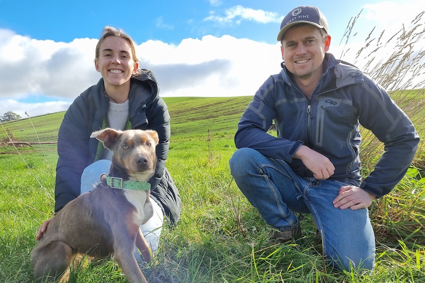 A young farming couple crouch with their dog on green grass, on a clear winter's day.