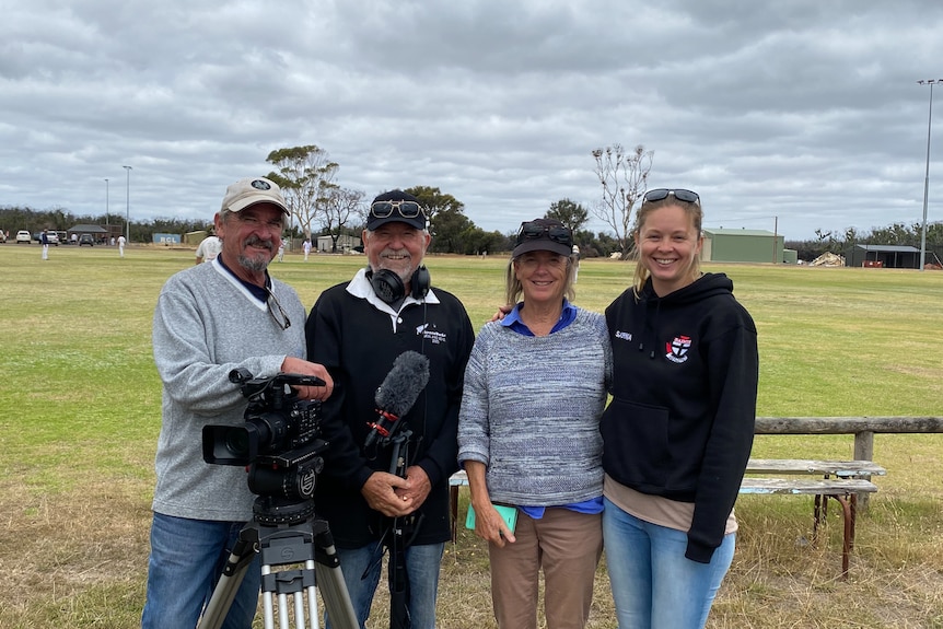 Two men and two ladies stand in front of a sporting field holding cameras and microphones