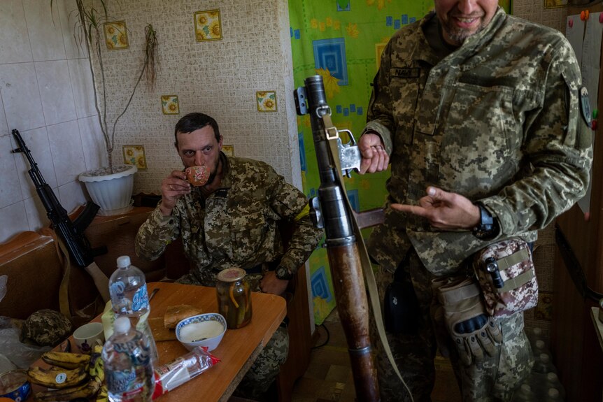 Two soldiers in a kitchen.  One drinks a cup of tea with his rifle propped up beside him. The other shows his weapon