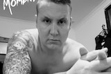 A black-and-white selfie-style shot of a shirtless man in his 30s with a sleeve tattoo.