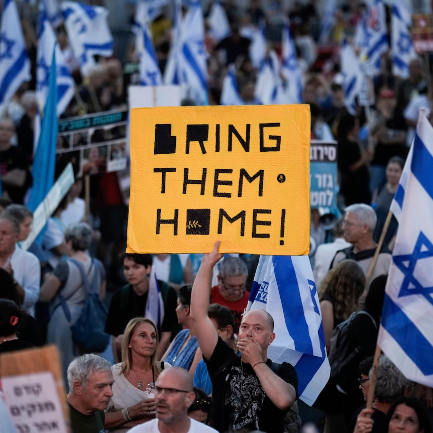 A man stands in a crowd of Israeli supporters holding a yellow and black signs with the words 'Bring them Home' on it