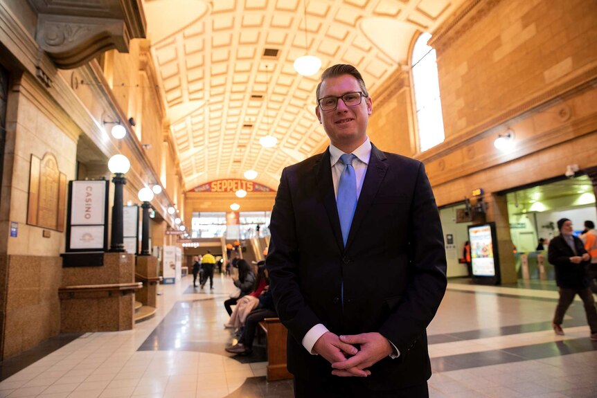 A bespectacled middle-aged man wearing a dark suit and a powder-blue tie stands in a transport interchange of some kind.