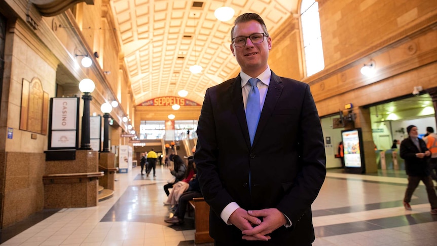 A bespectacled middle-aged man wearing a dark suit and a powder-blue tie stands in a transport interchange of some kind.