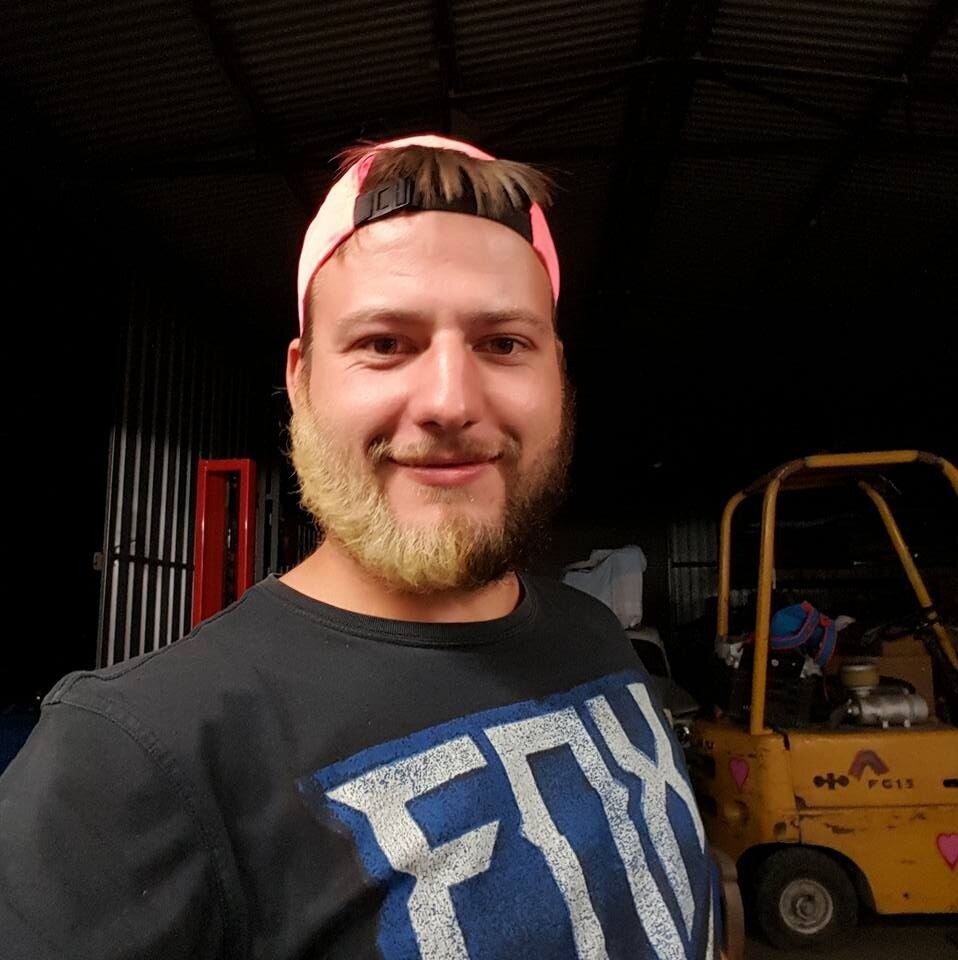 A man wearing a baseball cap and a brown beard, half of the beard is bleached blonde 