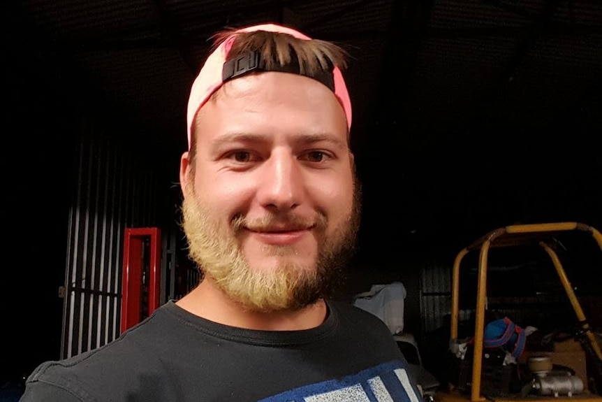 A man wearing a baseball cap and a brown beard, half of the beard is bleached blonde 