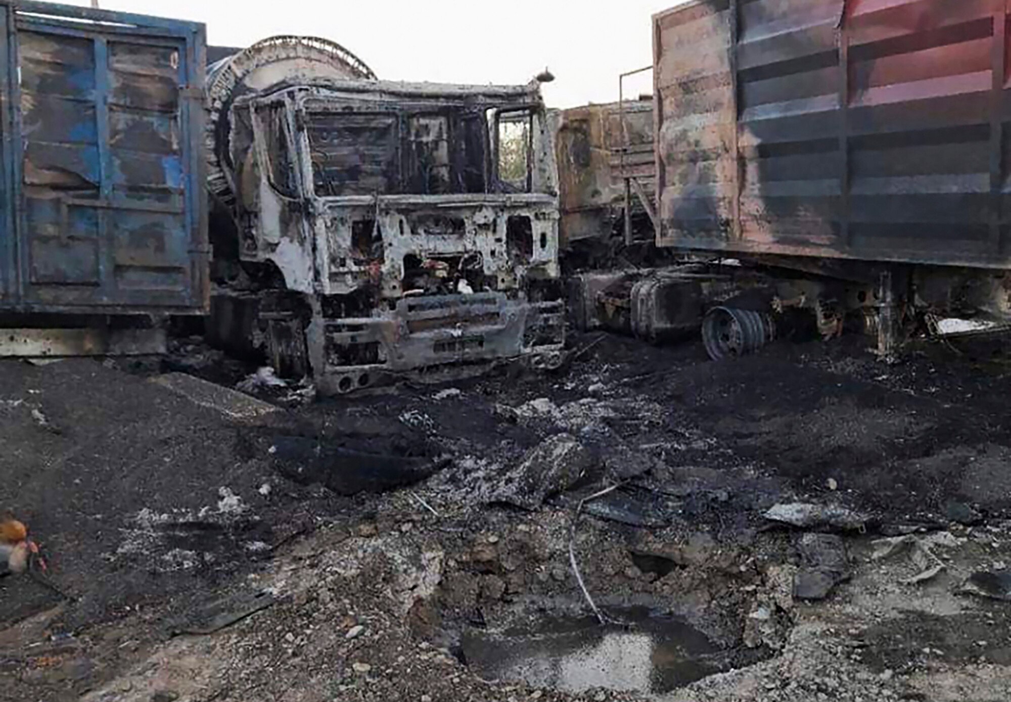 A medium shot of destroyed and burnt out trucks, next to a damaged shipping container and piles of dirt and rubble.
