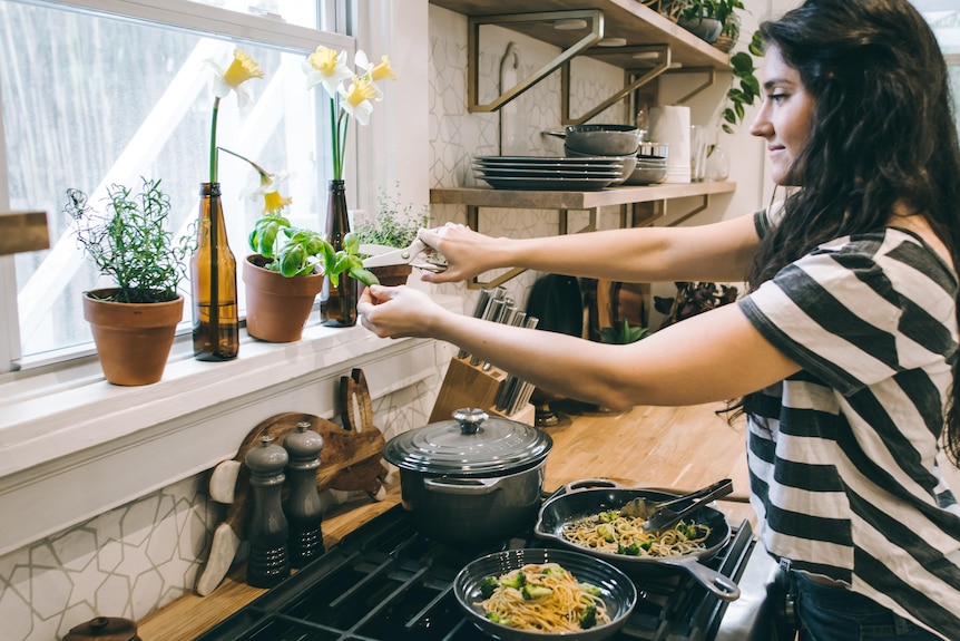 Woman cooking in saucepans at stove and reaching for basil leaf.