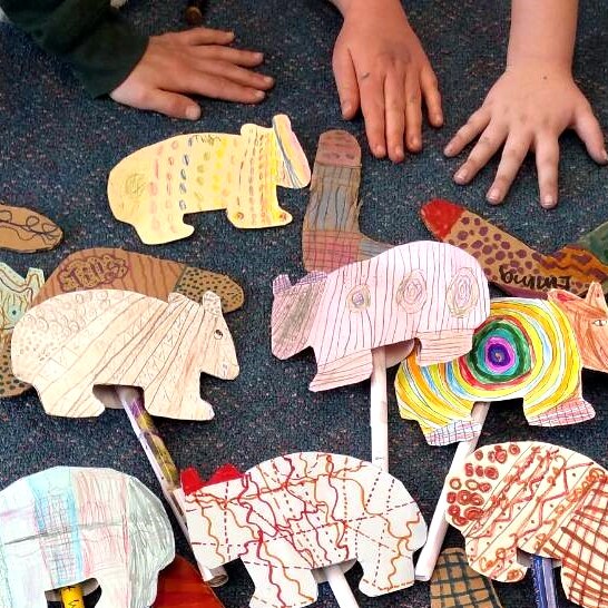 A collection of coloured cardboard wombats and boomerangs on a carpet with children's hands.
