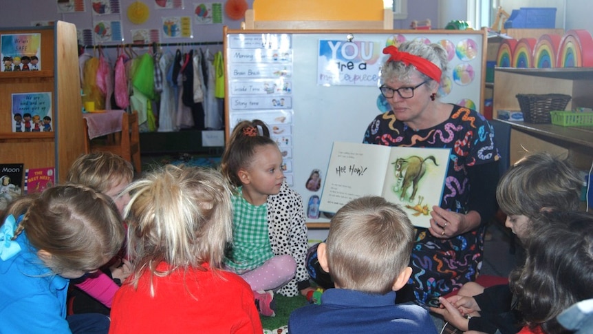 a woman sits on a chair reading a book, to a small group of young kids.