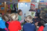 a woman sits on a chair reading a book, to a small group of young kids.