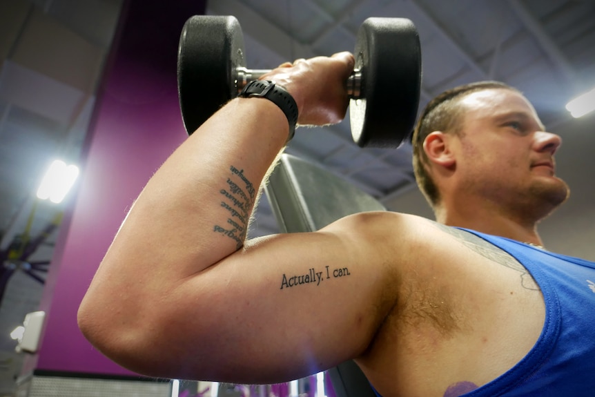 A man lifts a weight showing a bicep tattoo that reads 'Actually I can'