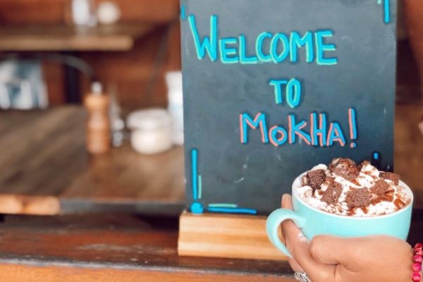 A hand holds a coffee in front of a sign that says: "Welcome to Mokha".