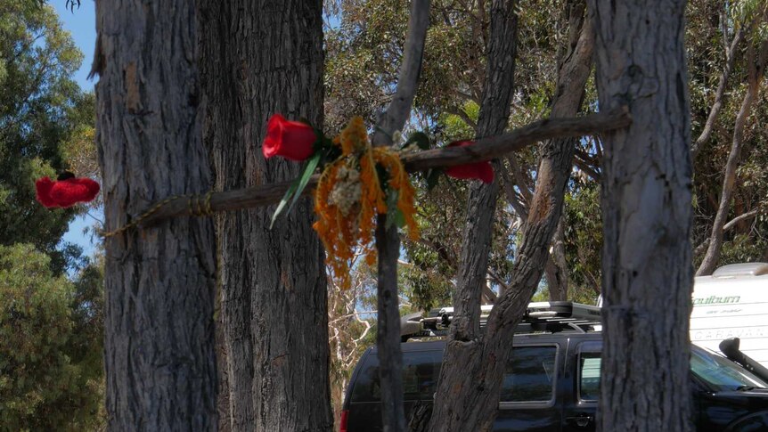 A cross made of sticks with flowers on it and flowers at the bottom. A black car is parked in the background.
