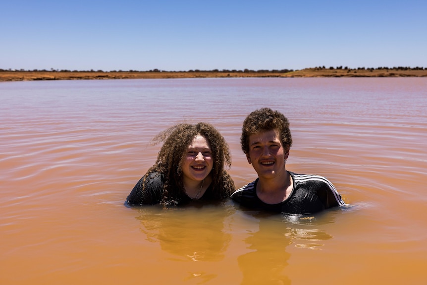 Young man and woman sit in the brown water of a dam, smiling.