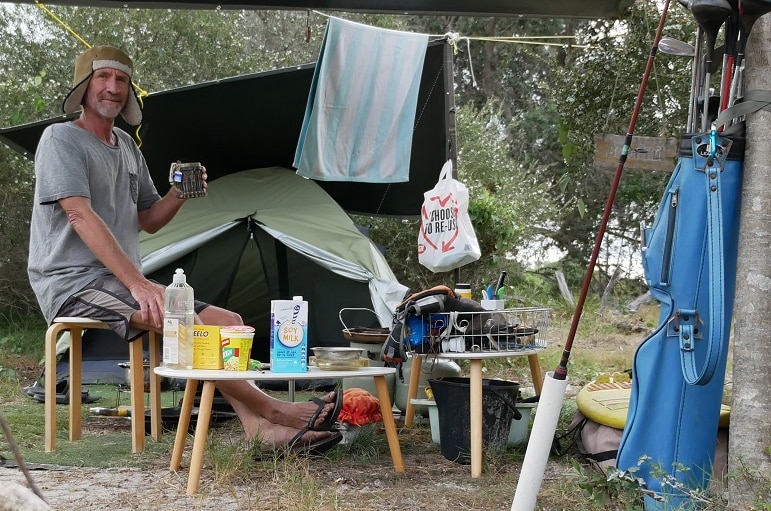 A man holds a mug of tea as he sits on a stool under a suspended tarpaulin in front of a tent set up in the bush.