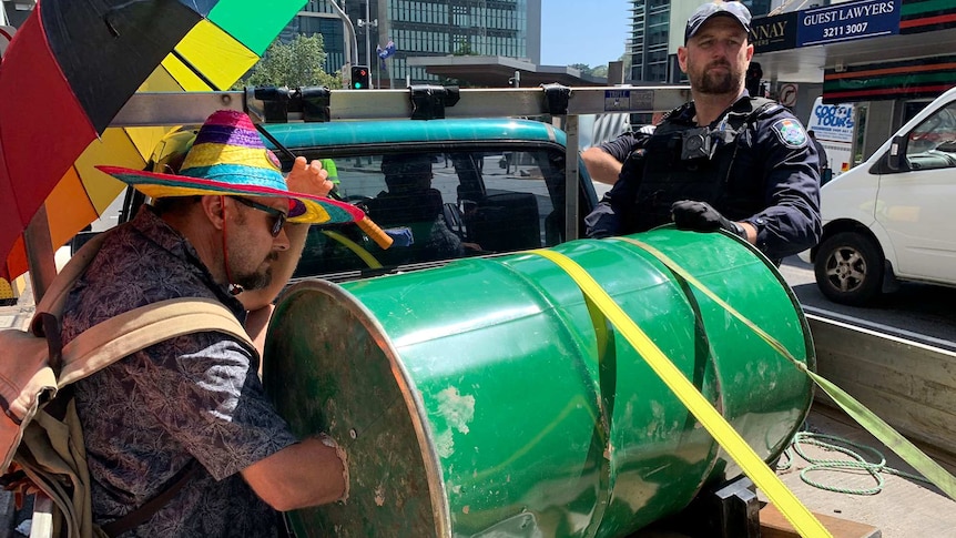 An Extinction Rebellion protester sits in the back of a ute with one arm in a barrel while the other holds an umbrella.