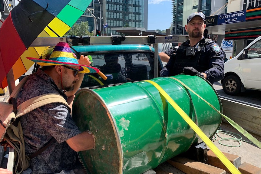 An Extinction Rebellion protester sits in the back of a ute with one arm in a barrel while the other holds an umbrella.