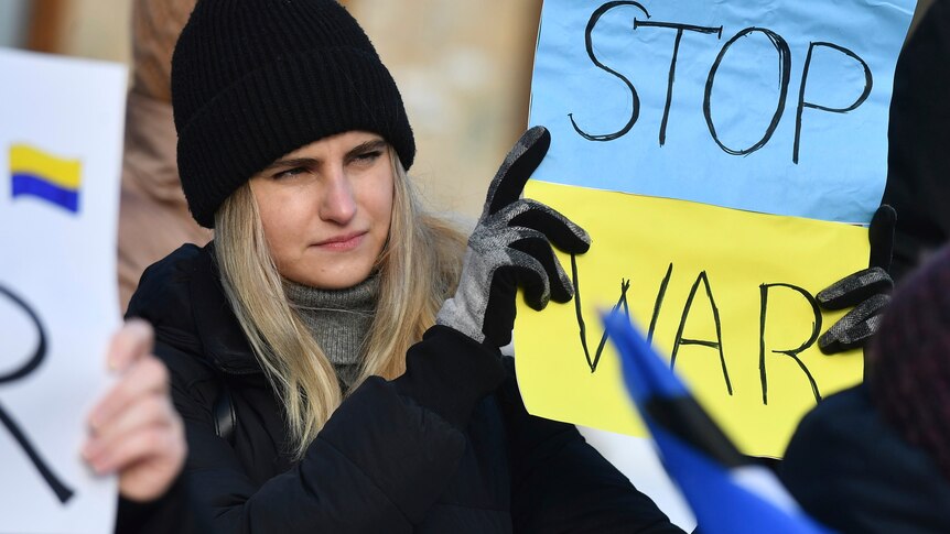 A woman wearing a black coat, beanie and gloves holds a blue and yellow sign that reads "stop war".
