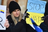 A woman wearing a black coat, beanie and gloves holds a blue and yellow sign that reads "stop war".