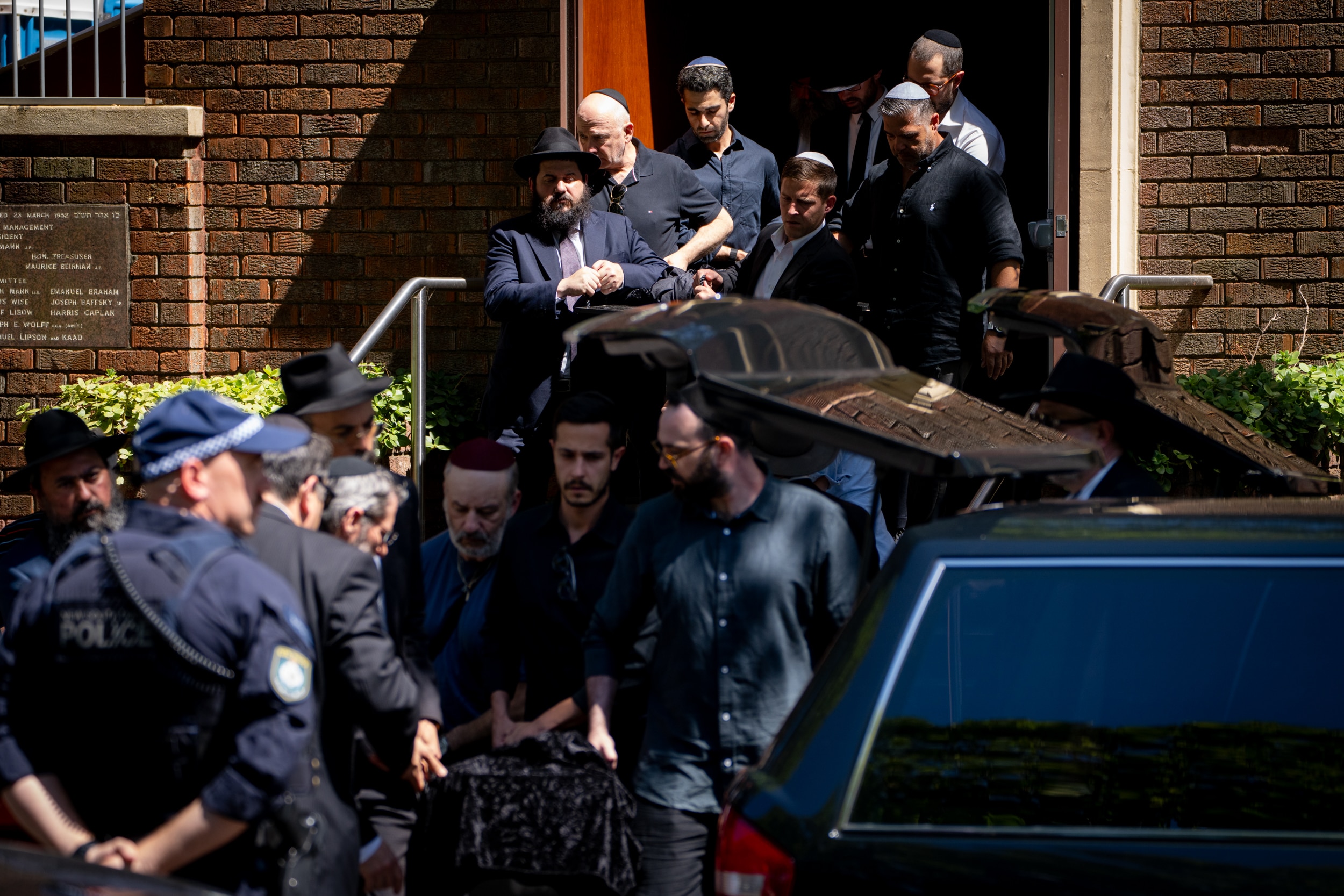 Un grupo de personas se reúne cerca de un coche fúnebre, con un oficial de policía junto a él.
