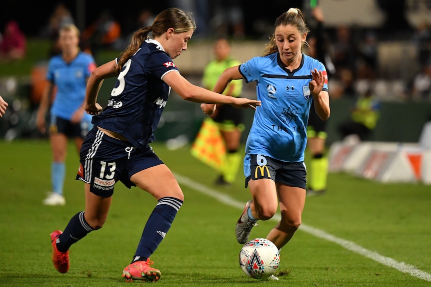 A Sydney FC W-League player dribbles the ball as a Melbourne Victory opponent runs alongside her.