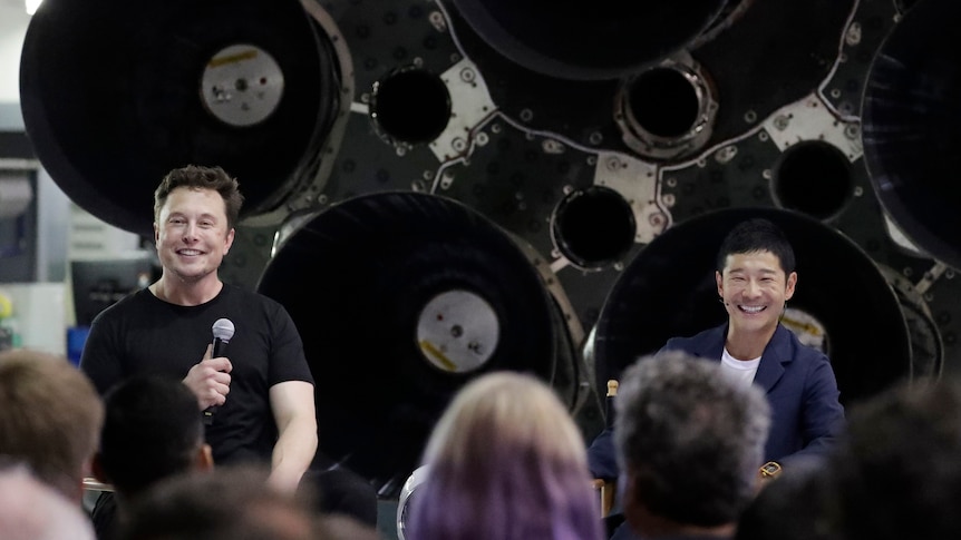 Two men sit smiling in front of what appears to be the underside of a rocket