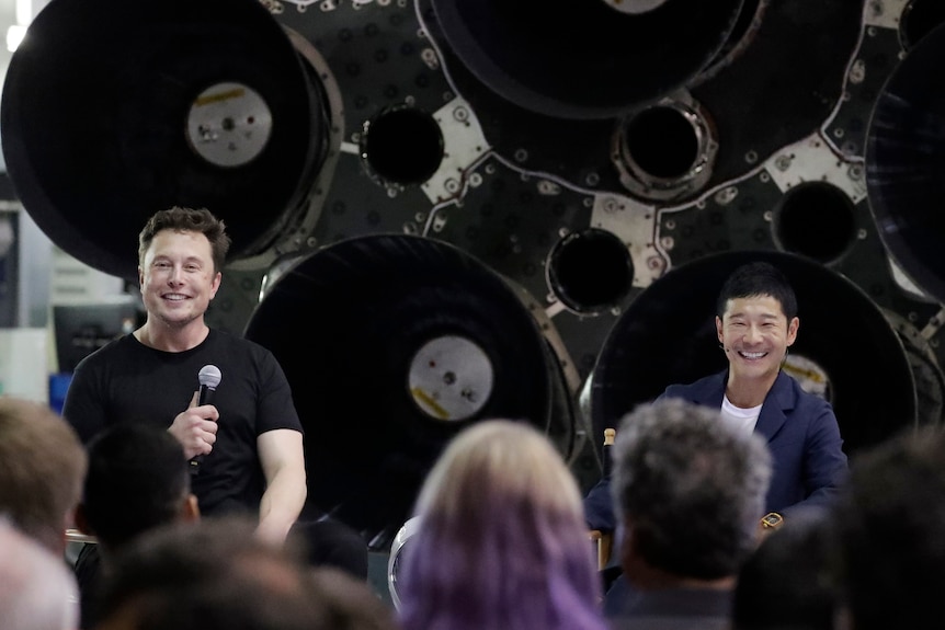 Two men sit smiling in front of what appears to be the underside of a rocket