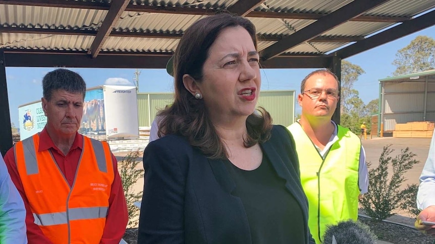 Annastacia Palaszczuk stands under a shed talking to the media with men wearing high viz vests on either side of her