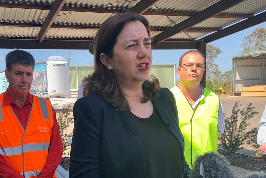 Annastacia Palaszczuk stands under a shed talking to the media with men wearing high viz vests on either side of her
