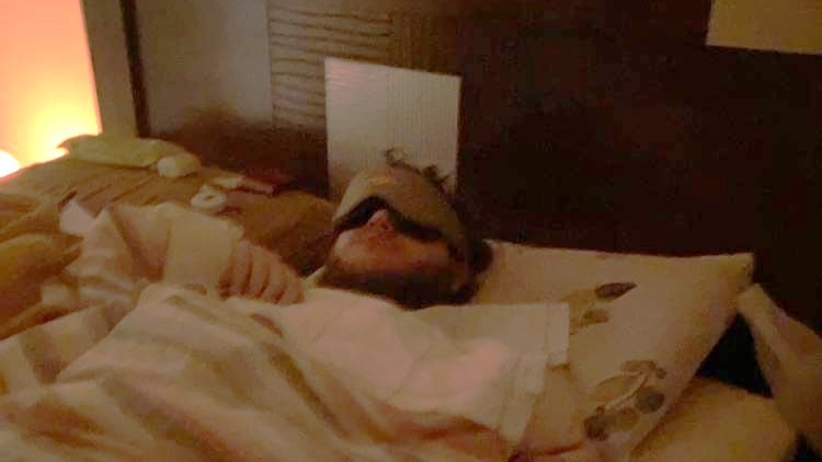 Man lying in bed with mask on, piece of paper on the bedhead, striped blanket, brown headrest 
