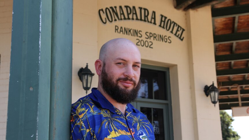 A bearded, bald man in a blue paramatta eels collared shirt poses in front of a country pub. 