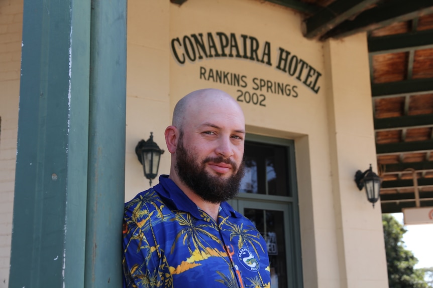A bearded, bald man in a blue paramatta eels collared shirt poses in front of a country pub. 
