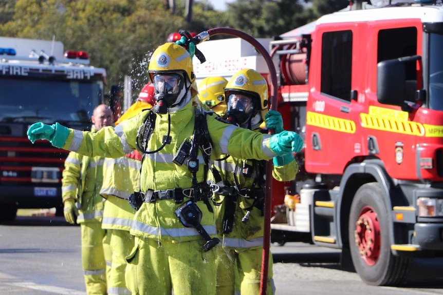 A firefighter in full protective clothing and gas mask is hosed down with water by another firefighter.