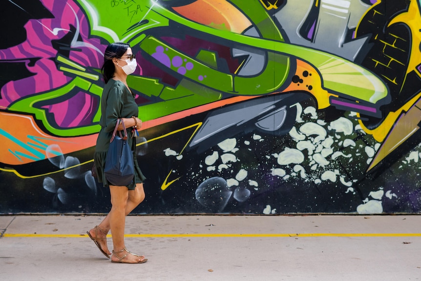 Une femme portant un masque facial marche à côté de graffitis colorés dans le CBD de Darwin.