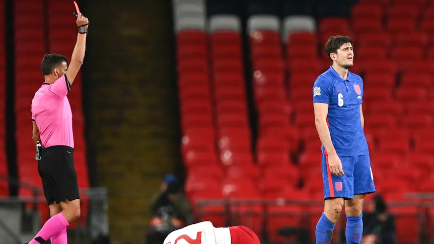 A footballer looks upset as a referee holds red card — meanwhile an injured player lies on the turf.