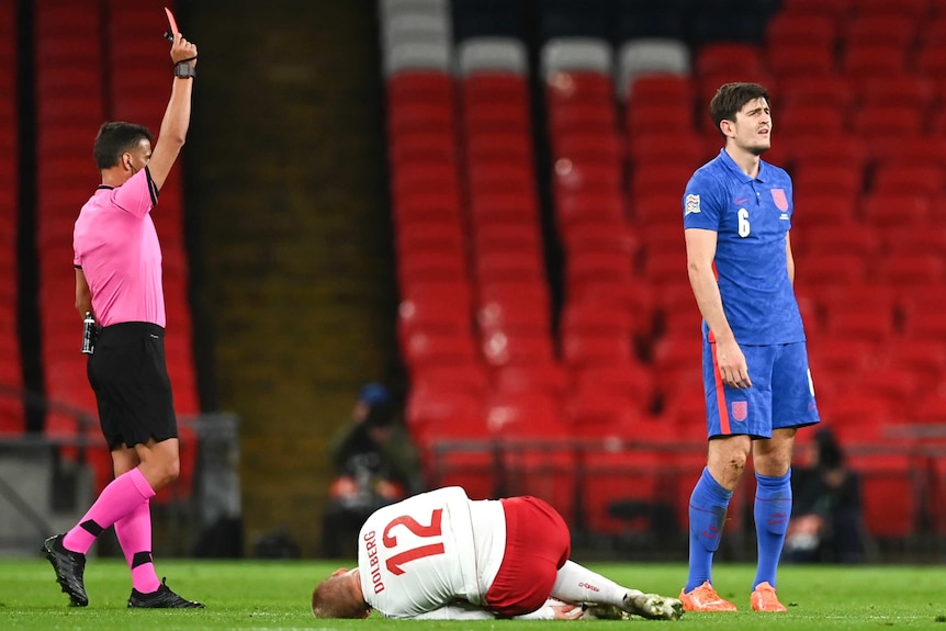 A footballer looks upset as a referee holds red card — meanwhile an injured player lies on the turf.