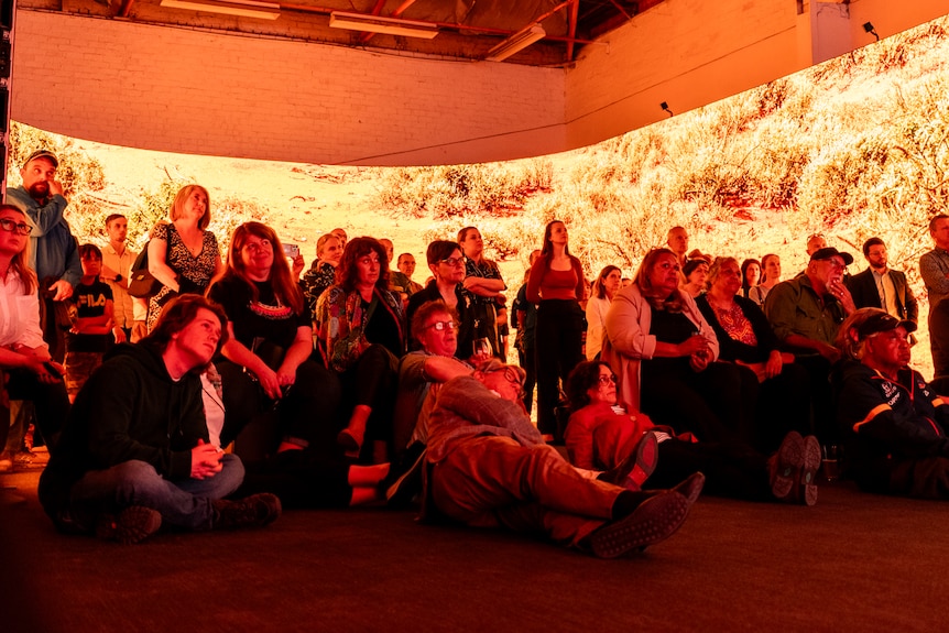A small crowd of people sit and stand in darkness inside a curved LED screen showing orange images of bush scrub. 