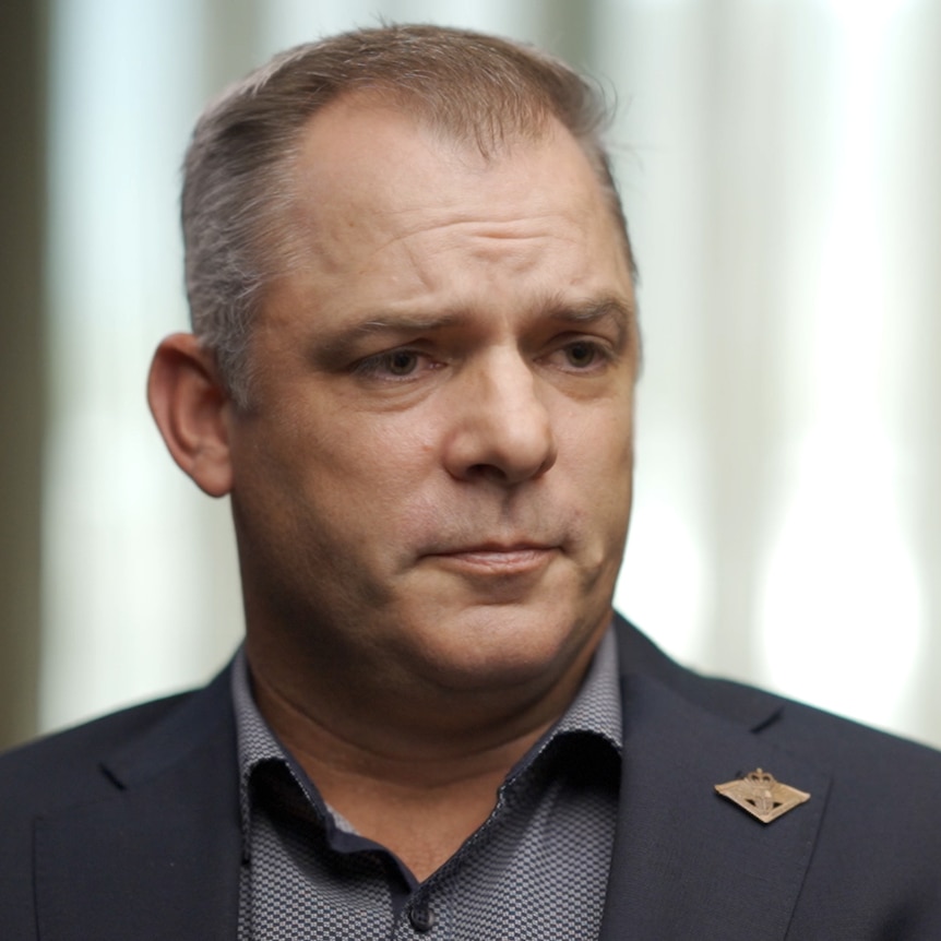 A man wearing sitting indoors looks to the side. He wears a suit jacket with a pin on the lapel.