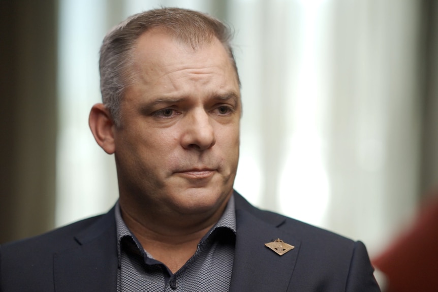 A man wearing sitting indoors looks to the side. He wears a suit jacket with a pin on the lapel.