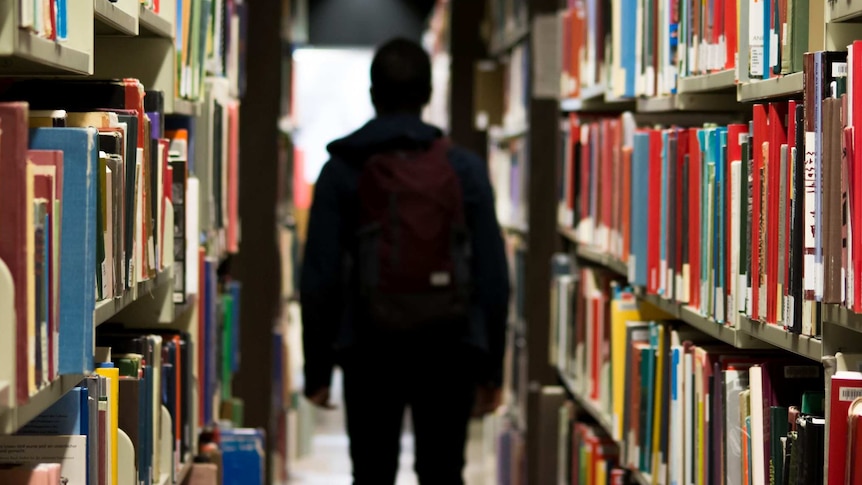 A student holding a backpack stands in shadow in a school library.