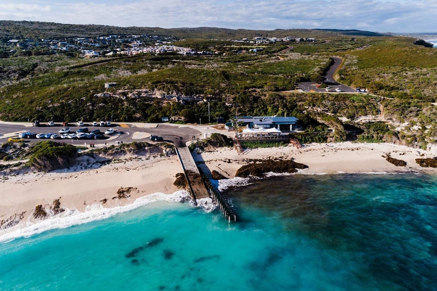 An aerial shot of a beach with a jetty and carparks in the foreground and native bushland in the background.
