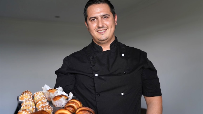 A chef in uniform holds a rack of pastries while smiling at the camera.