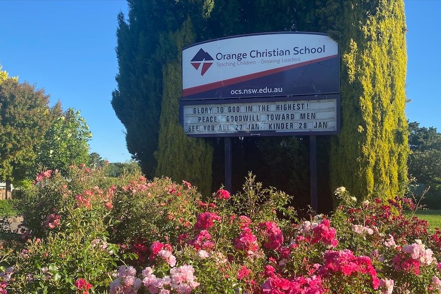 A school sign reads Orange Christian School behind a row of pink rose bushes.
