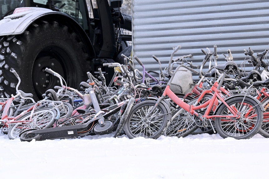 Dozens of bikes lighlty covered in snow lie on the ground next to a building