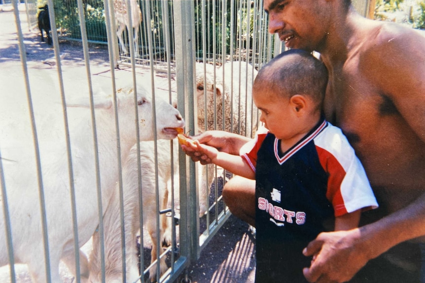 A man holds the hand of a boy who is feeding a goat through the bars of a metal fence.