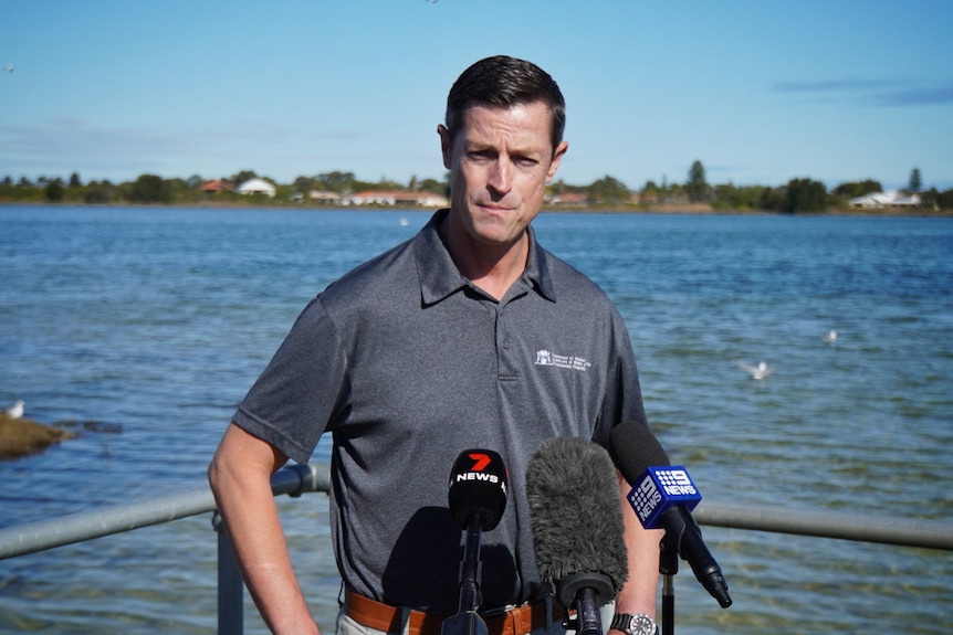 A man with short dark hair stands in front of water while addressing media