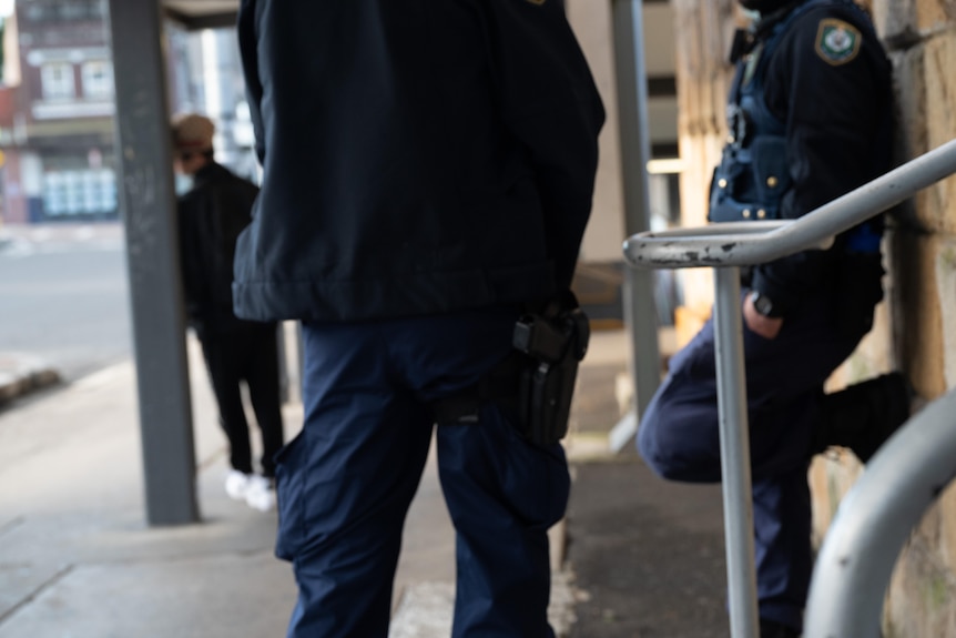 Two anonymous police officers stand and lean against a wall. In front of them leaning against a pole is a teen boy wearing a cap